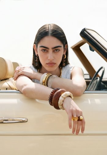 Femme aux cheveux foncés et bijoux audacieux reposant ses bras sur la porte d’une voiture décapotable beige, regardant droit devant avec une expression neutre.