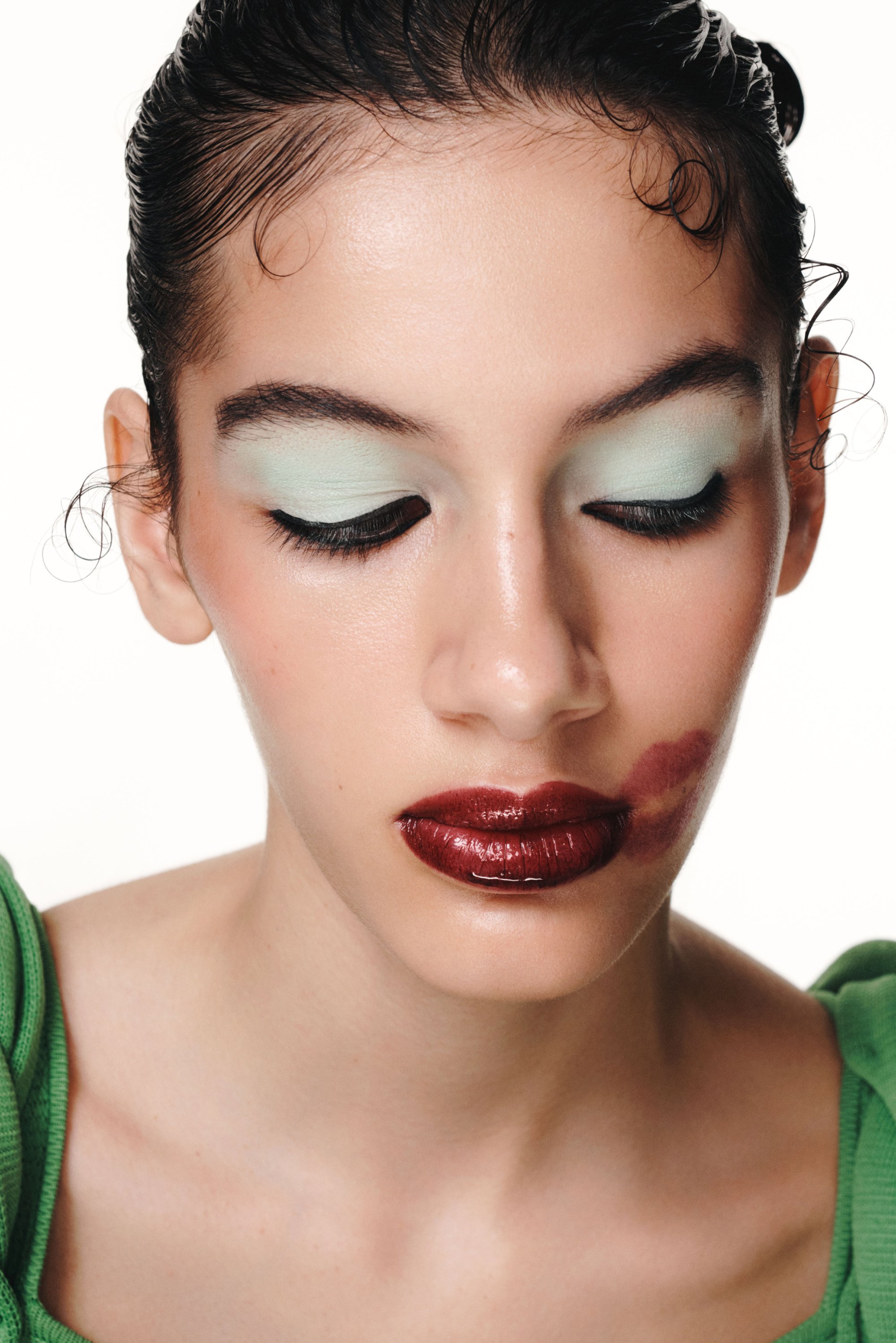 Una modelo con el cabello estilizado muestra un maquillaje audaz: sombra de ojos verde menta, labios rojos oscuros y brillantes, y un contorno de labios difuminado, vistiendo una blusa verde.