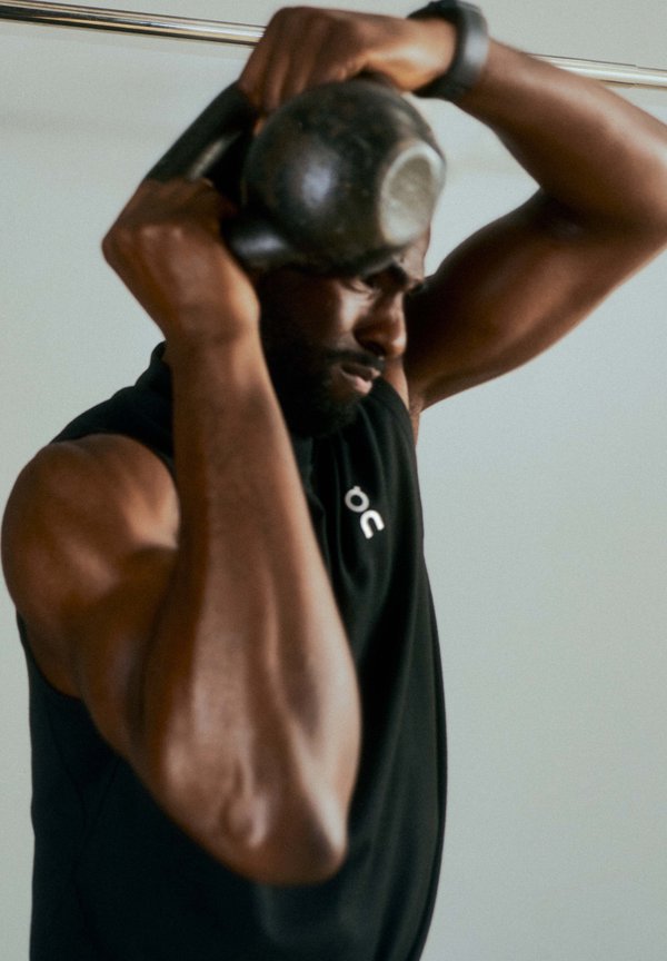 Muscular man in black sleeveless shirt lifting a kettlebell overhead, focusing on strength training indoors.