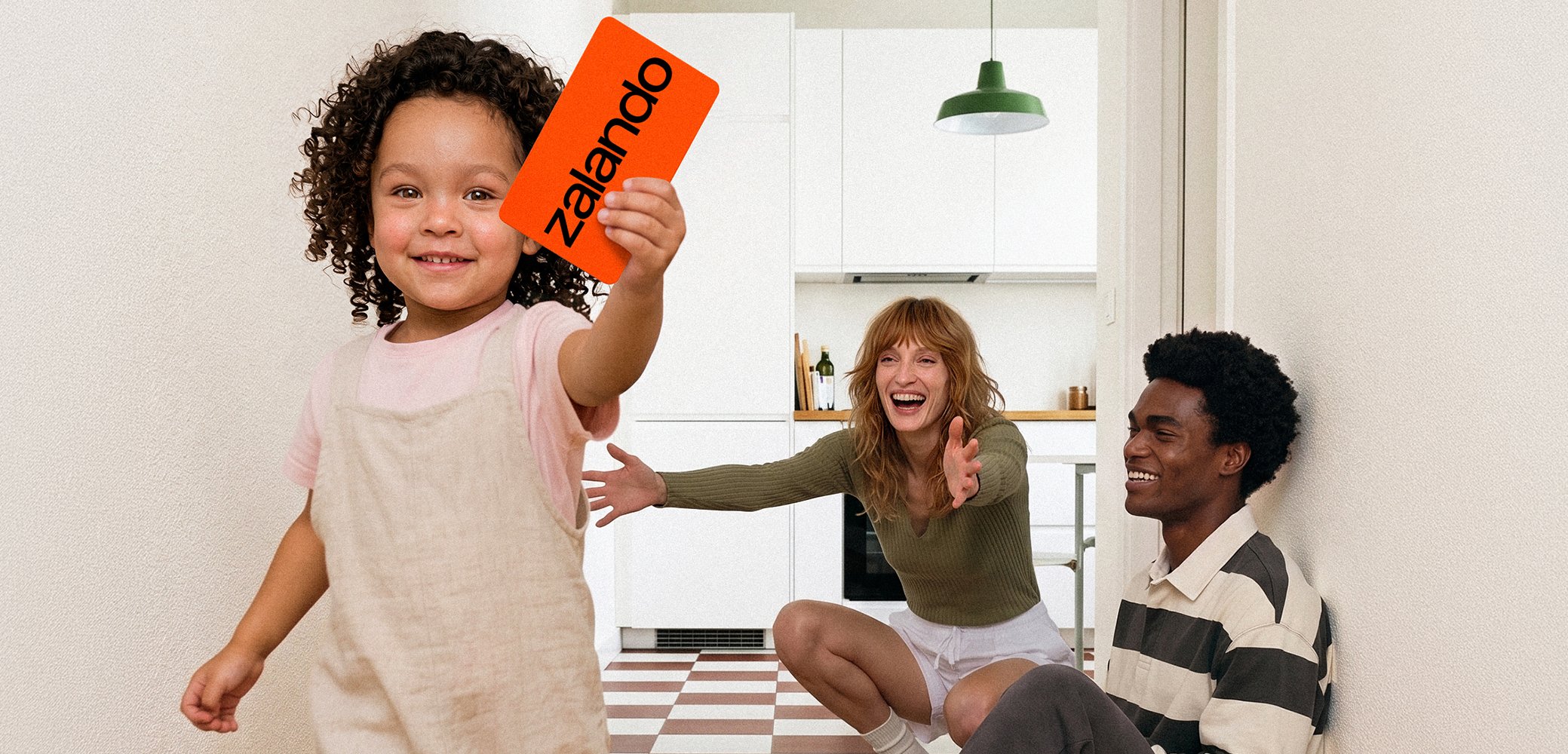 Un niño sostiene una tarjeta naranja de Zalando hacia la cámara mientras una mujer y un hombre sonrientes están sentados en el suelo de una cocina alcanzando al niño.
