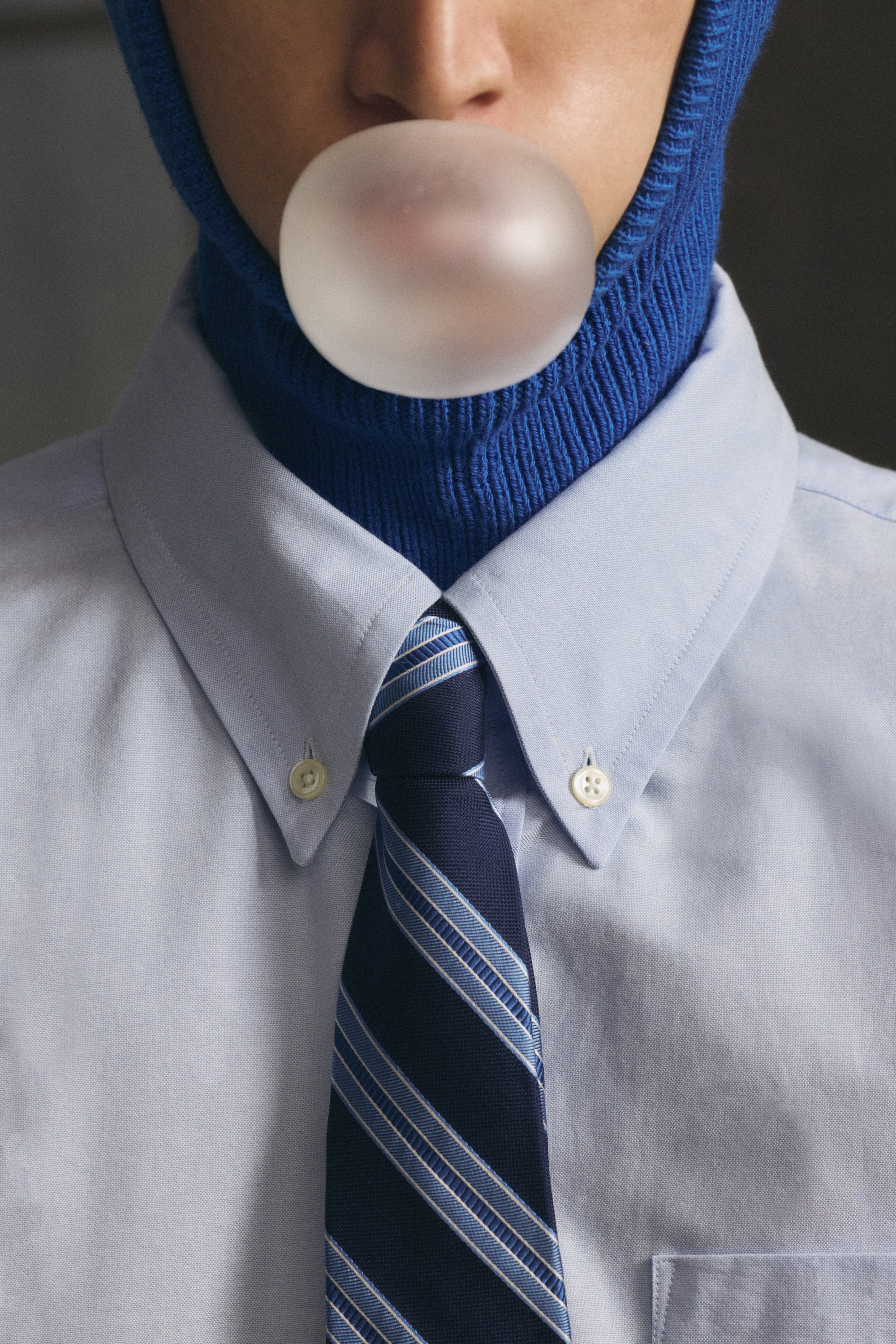 En person har på sig en blå stickad mössa och en ljusblå skjorta med en randig slips, och blåser en stor bubbla med tuggummi.