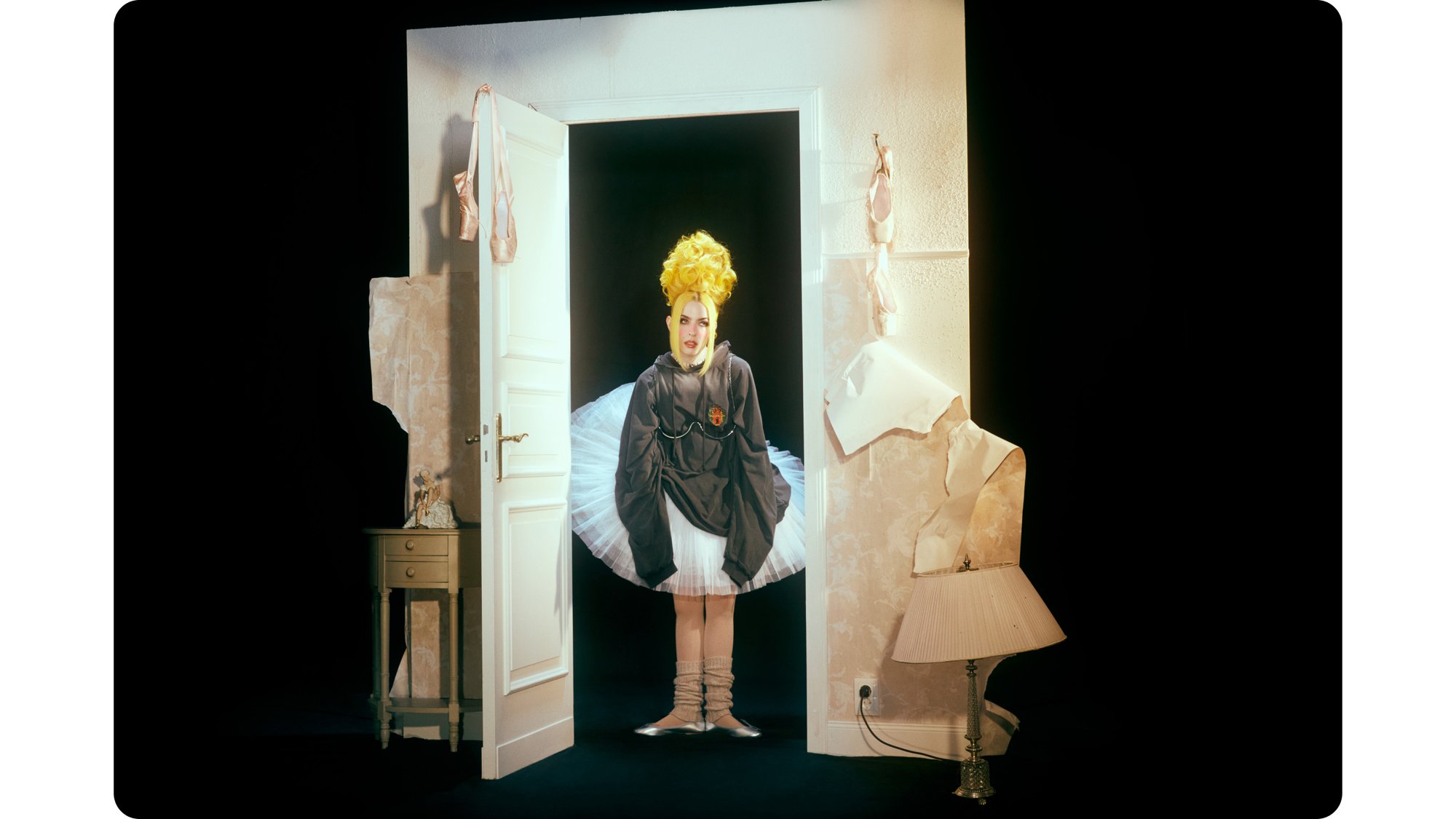 A person with a large yellow hairstyle wears a gray outfit over a tutu, standing in a doorway decorated with ballet shoes and wallpaper scraps.