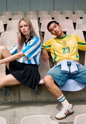 Eine junge Frau in einem blau-weiß gestreiften Hemd und schwarzem Rock sitzt neben einem jungen Mann in einem gelben Brasilien-Trikot und Jeans-Shorts auf Stadionplätzen.