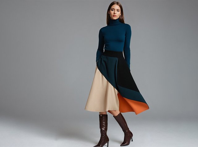 Woman wearing a blue turtleneck, color-blocked beige, black, teal, and orange skirt, and knee-high brown heeled boots, standing against gray background.