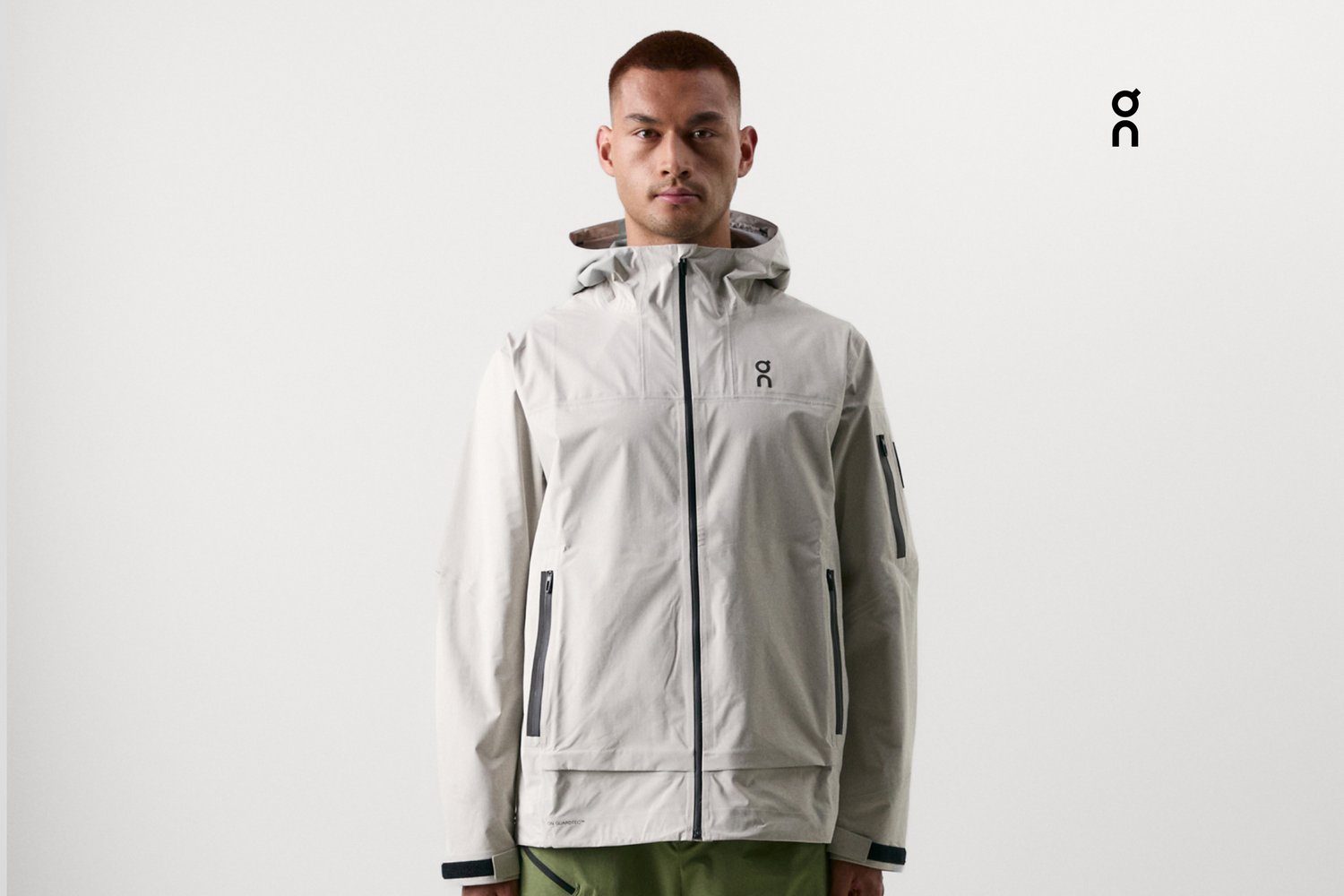 A young man wearing a light gray jacket with a hood and zippered pockets stands against a white background, appearing confident and poised.