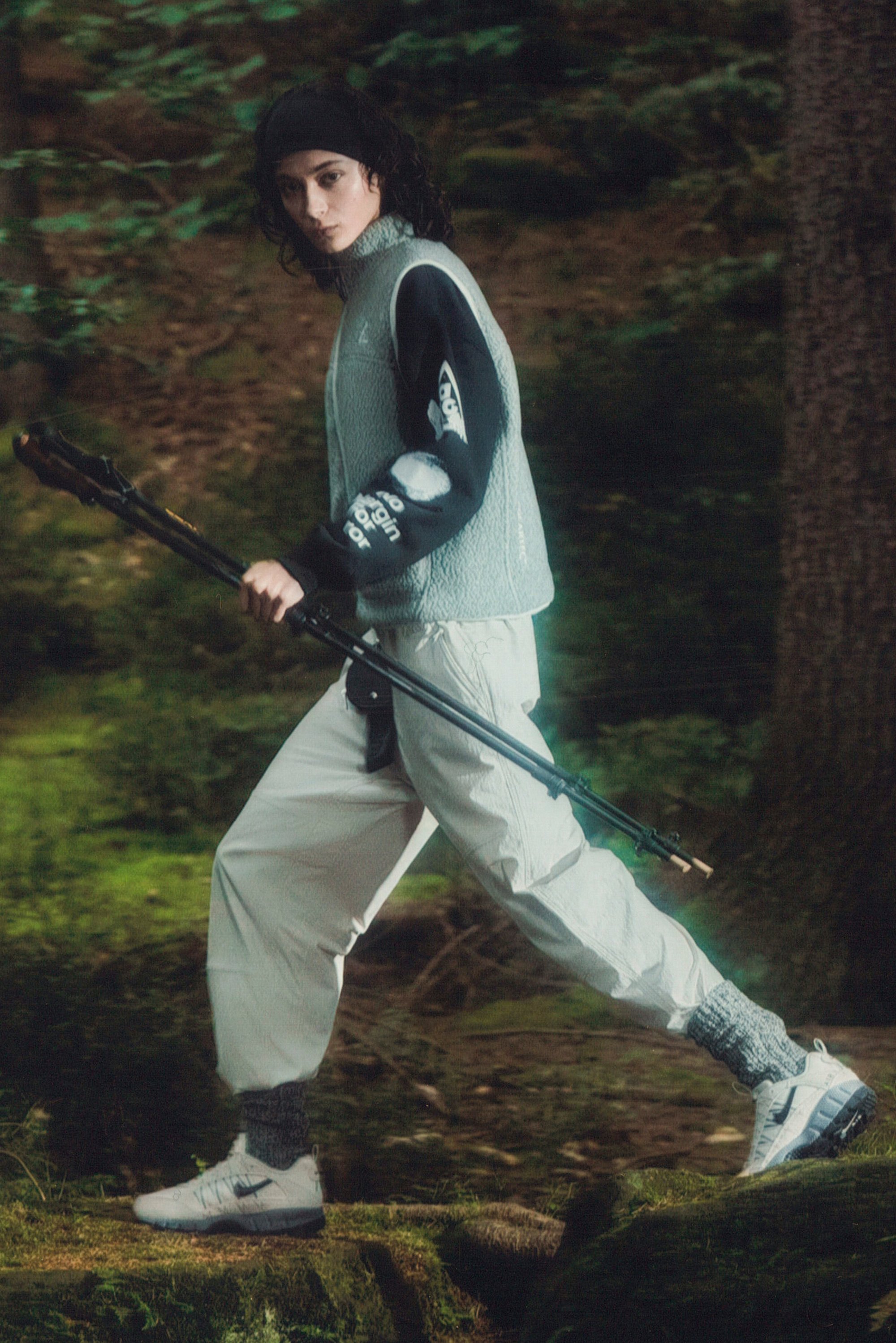 Una persona camina con confianza a través de un bosque, vistiendo un chaleco gris, mangas negras y pantalones blancos, sosteniendo bastones de senderismo.