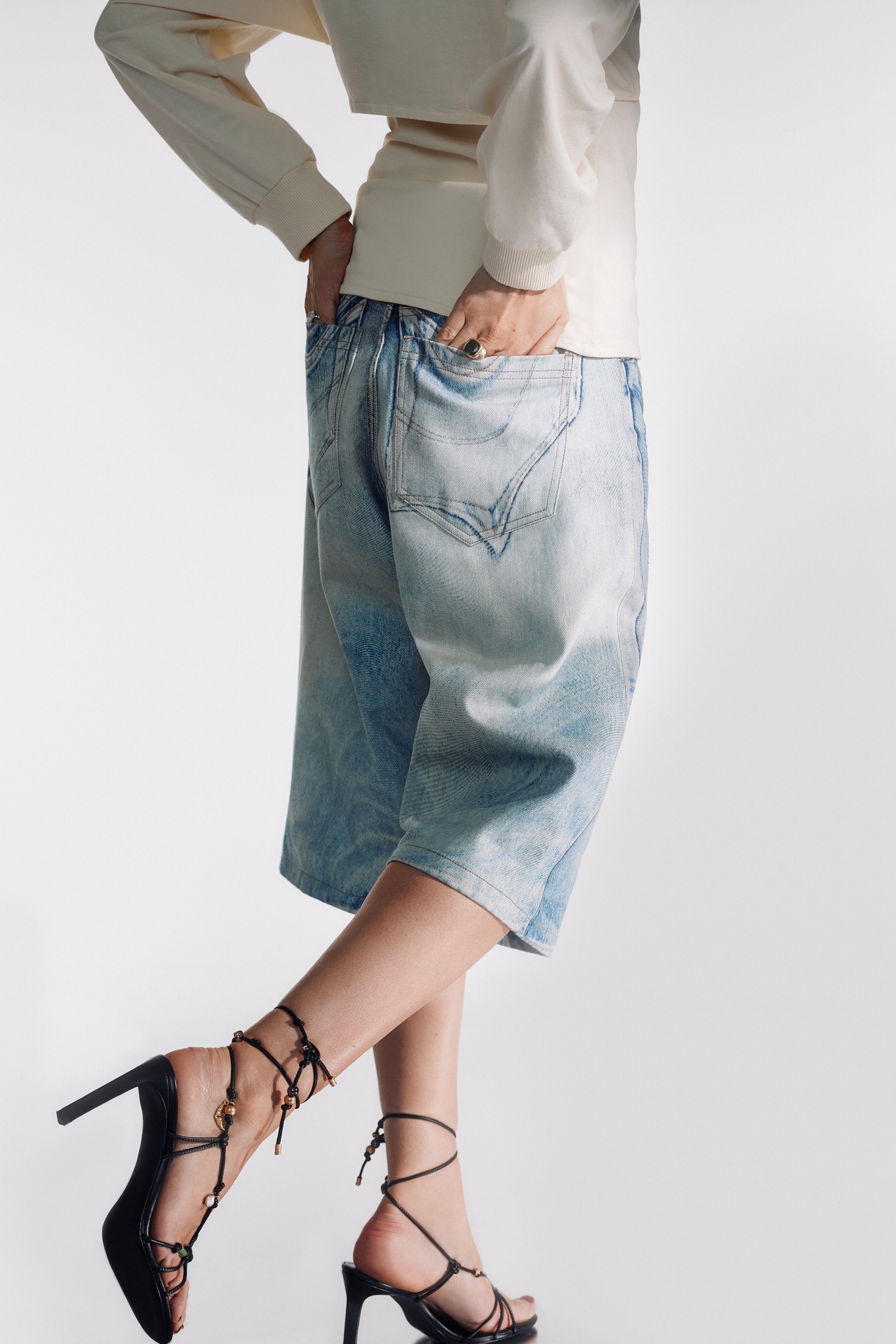 Una persona lleva una blusa corta color crema y pantalones cortos de mezclilla azul claros, mostrando unas sandalias de tacón negro con tiras y cuentas decorativas.