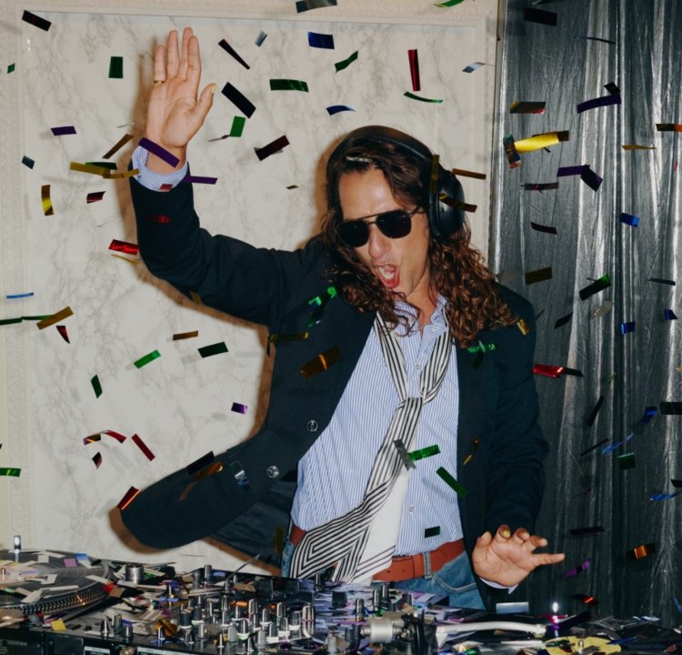 DJ-bås med skivspelare och färgglatt konfetti som regnar ner. DJ:n bär svarta solglasögon, en blazer och en randig slips.