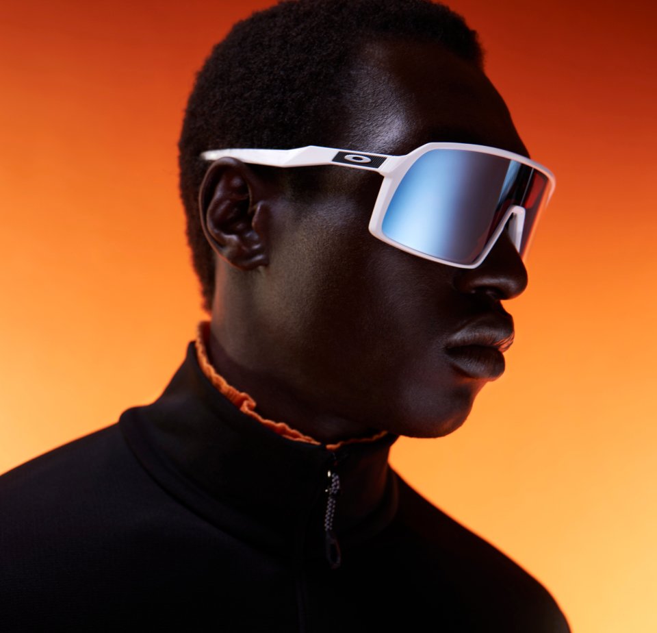 Person wearing futuristic white sunglasses with mirrored lenses, facing right, against a vibrant orange background, in a black high-collared top.