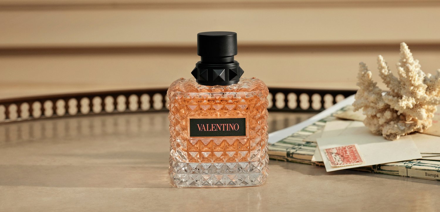 Doorzichtige glazen Valentino parfumfles met oranje vloeistof en zwarte dop op een tafel naast vintage brieven en koraaldecoratie.