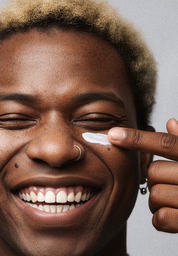 Persona sonriente aplicando crema blanca en la mejilla con el dedo, mostrando un pendiente en la nariz, cabello rubio corto y texturizado, y piel clara.