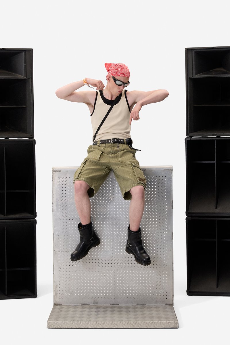 Ung mand iført solbriller, rødt pandebånd, tanktop, cargoshorts og sorte støvler, siddende på en metalplatform mellem to store sorte højttalere.