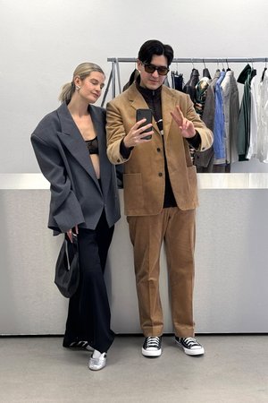 Deux individus élégants prennent un selfie dans un magasin de vêtements moderne. La femme porte un blazer surdimensionné et un pantalon noir élégant, tandis que l'homme arbore un costume en velours côtelé.