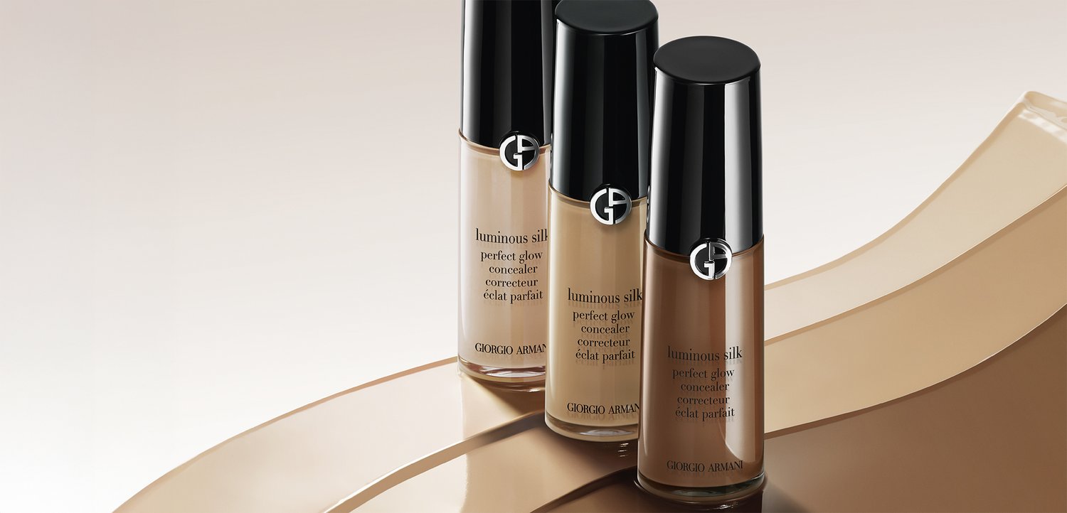 Drei Giorgio Armani Luminous Silk Perfect Glow Concealer-Flaschen in verschiedenen Hauttönen auf einer beige-farbenen, geschwungenen Oberfläche.