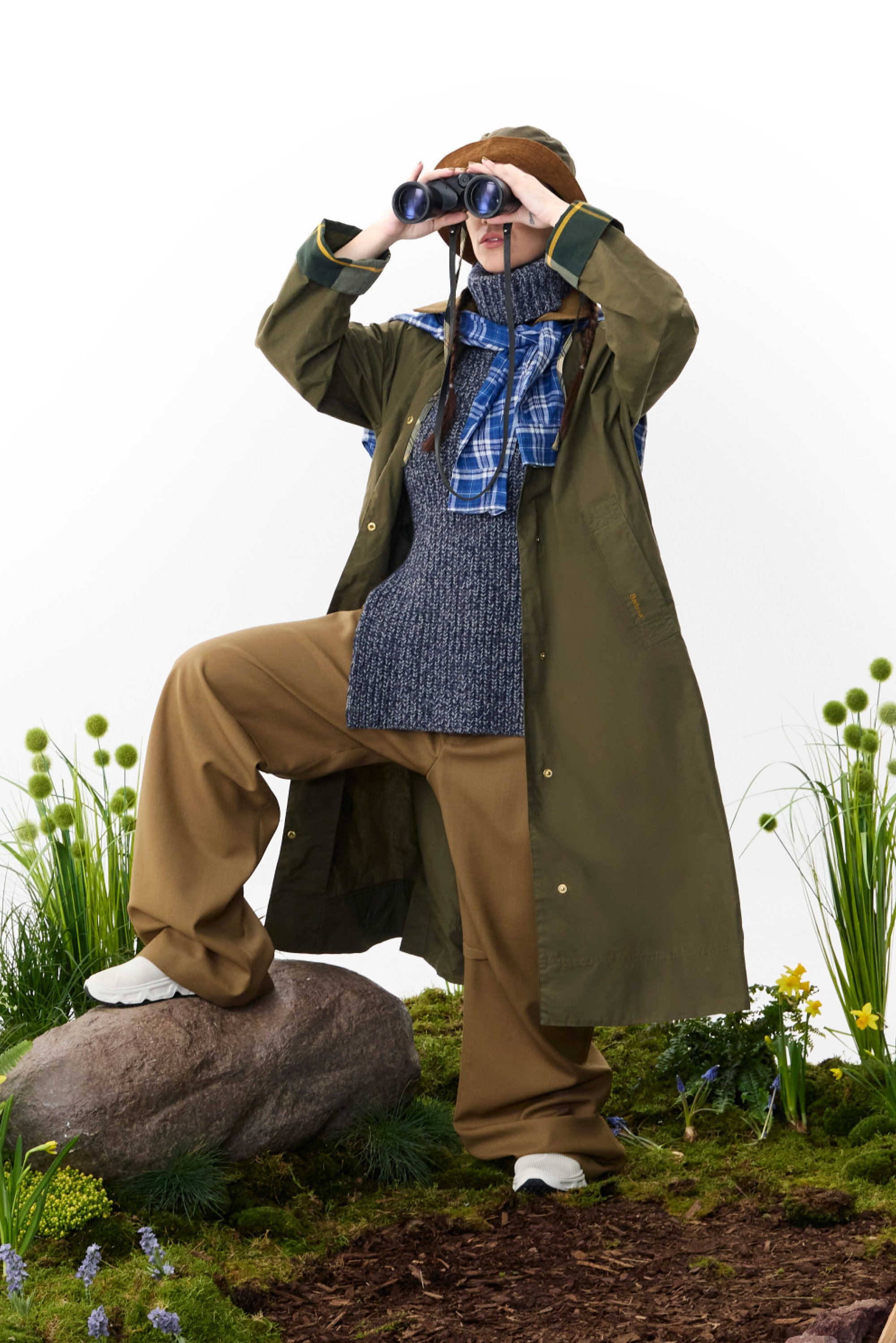 A person wearing a brown hat and green coat stands on a rock, holding binoculars and dressed in layered outdoor attire, surrounded by greenery.