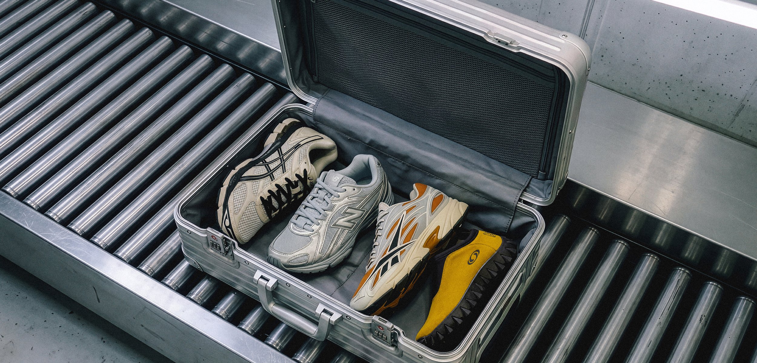 Quatre paires de baskets de styles et couleurs différents placées à l'intérieur d'une valise rigide argentée ouverte sur un tapis roulant à bagages.