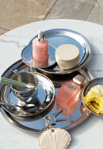 Zwei silberne Tabletts auf einem weißen Marmortisch halten Hautpflegeprodukte, eine Parfümflasche, eine Tasse mit Untertasse und einer grünen Mascara, Schlüssel mit einem Muschel-Schlüsselanhänger sowie ein teilweise gegessenes Gebäck.