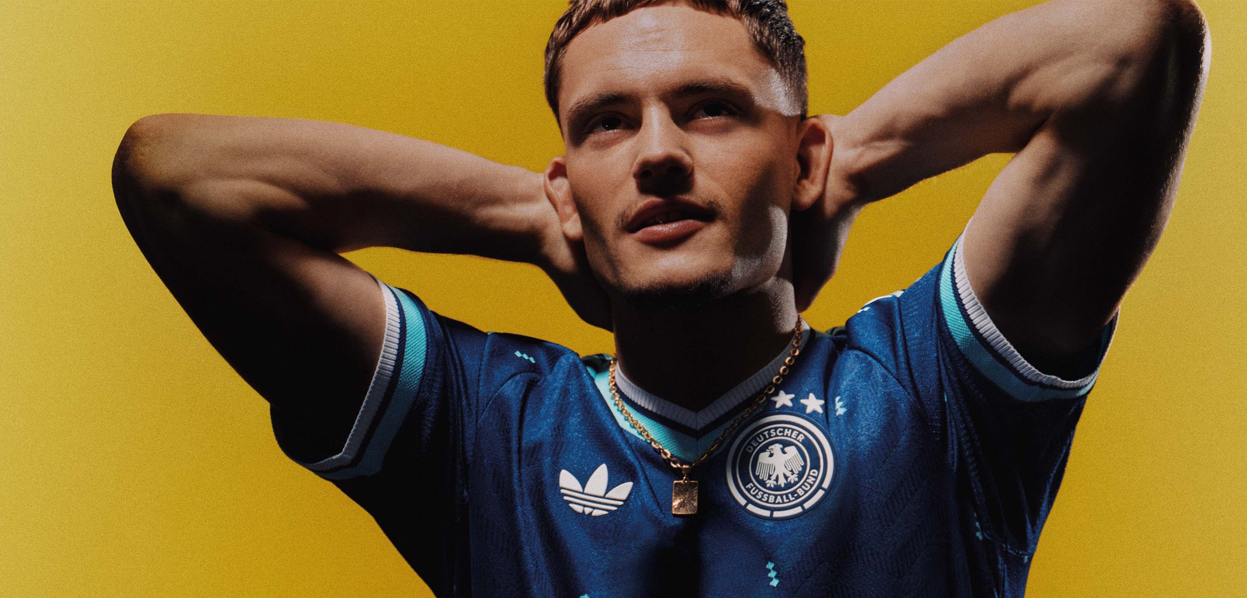 Jeune homme portant un maillot de football allemand bleu, posant avec les mains derrière la tête devant un fond jaune.