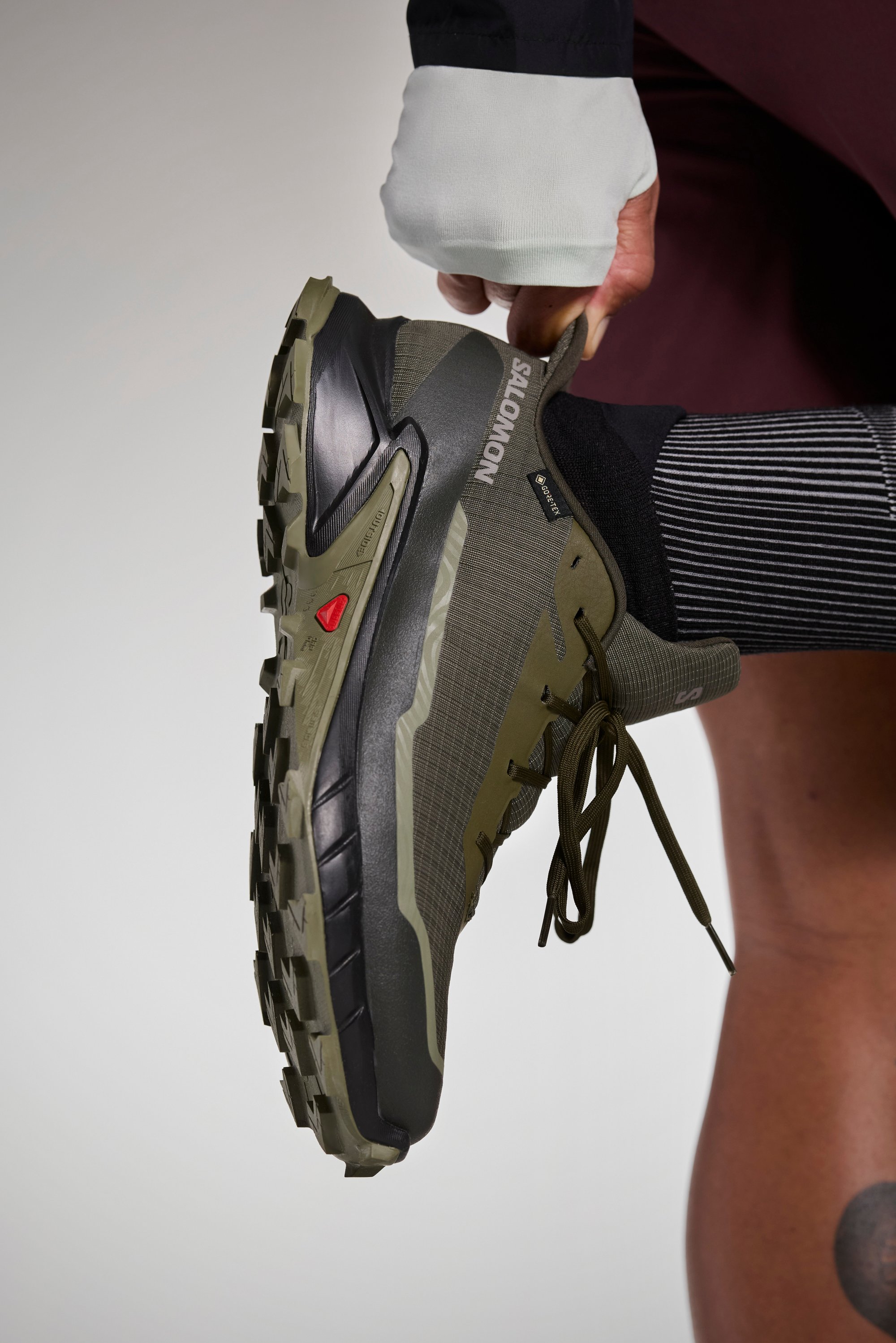 Una persona ajusta una zapatilla de trail running verde de Salomon, mostrando su suela texturizada y características, con un calcetín a rayas parcialmente visible.