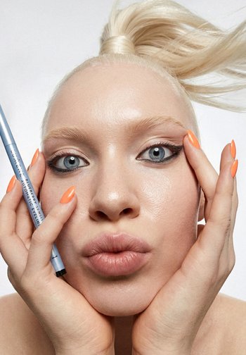 Close-up van een vrouw met oranje nagels die een blauwe eyelinerpotlood dicht bij haar gezicht houdt, met scherpe zwarte oogmake-up en naar achter getrokken blond haar.