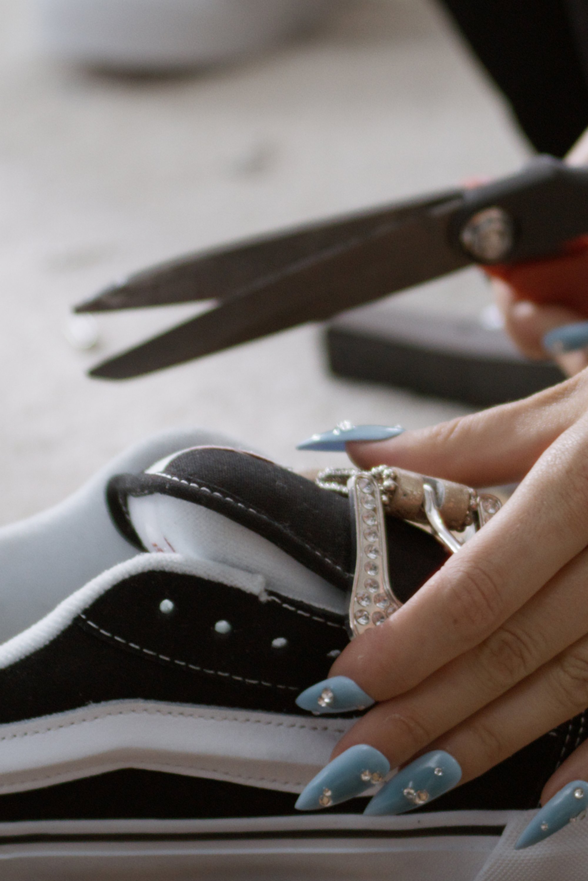 Una mano con largas uñas azules sostiene unas tijeras, recortando material en una zapatilla negra y blanca con correas decorativas.