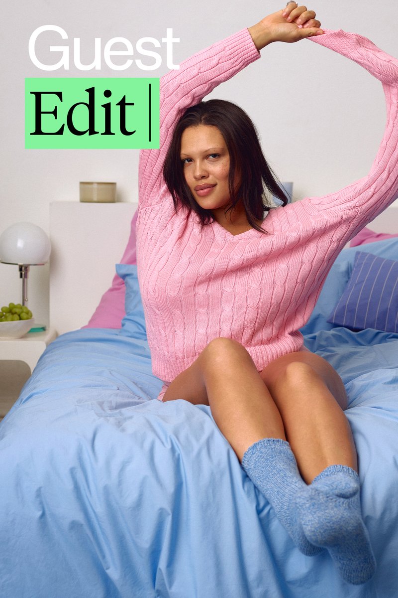 Una donna in un maglione rosa e biancheria intima è sdraiata su un letto con biancheria da letto blu e rosa. Un piccolo cane riposa accanto a lei. Il testo sovrapposto dice "Guest Edit."