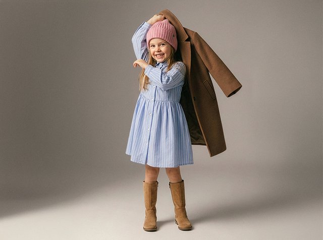 Jeune fille portant une robe bleue, un bonnet en tricot rose et des bottes brunes, tenant un manteau marron sur l'épaule, souriante et posant.
