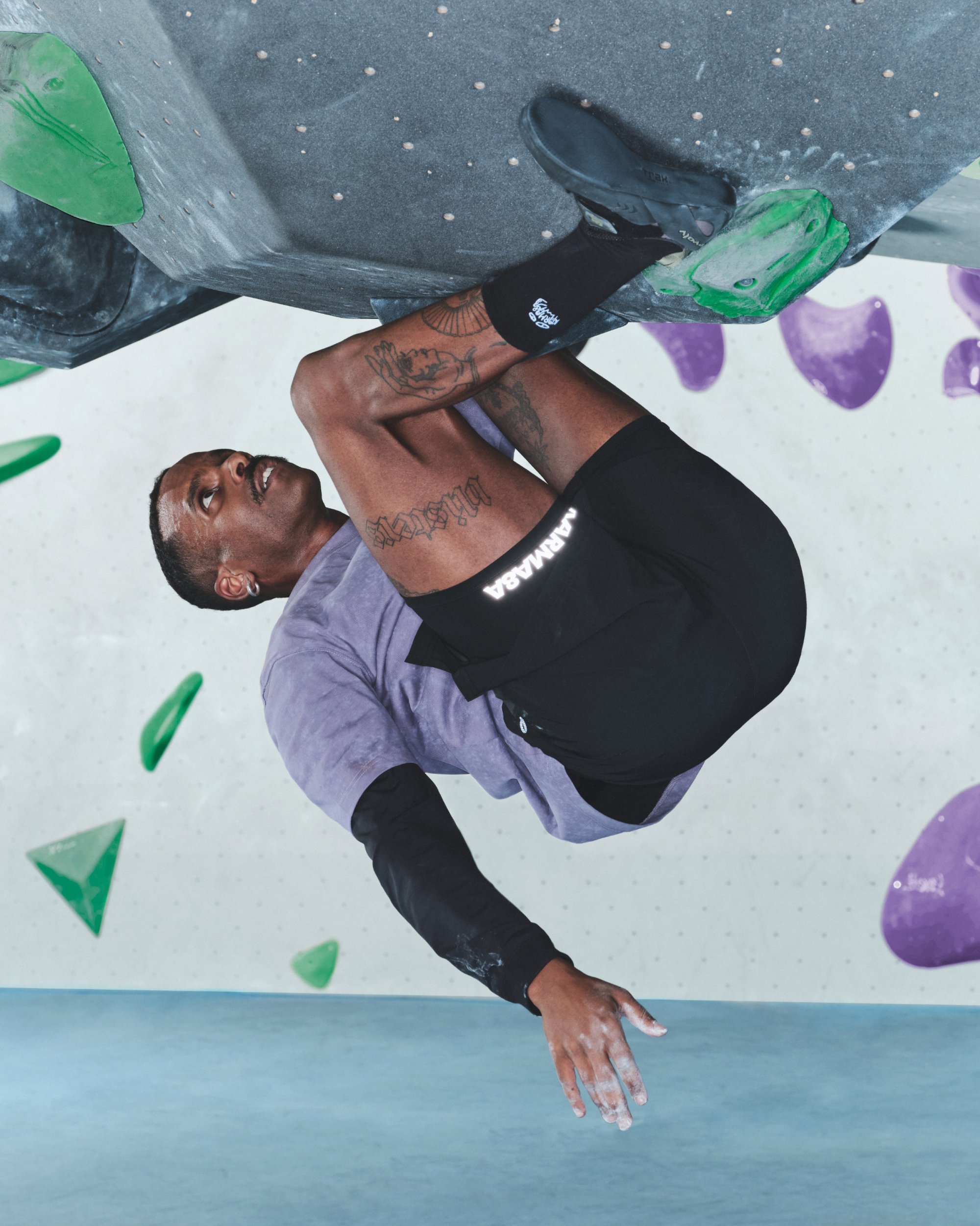 Escalador con una camiseta morada y pantalones cortos negros agarrándose de un agarre verde en una pared de escalada en bloque, con una superficie gris texturizada y agarres coloridos.