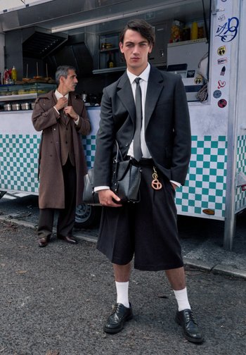 Schwarze Anzugjacke über einem weißen Hemd und einer schwarzen Krawatte; weite schwarze Shorts; schwarze Schuhe; hält eine strukturierte schwarze Tasche mit einem Brezel-Anhänger.