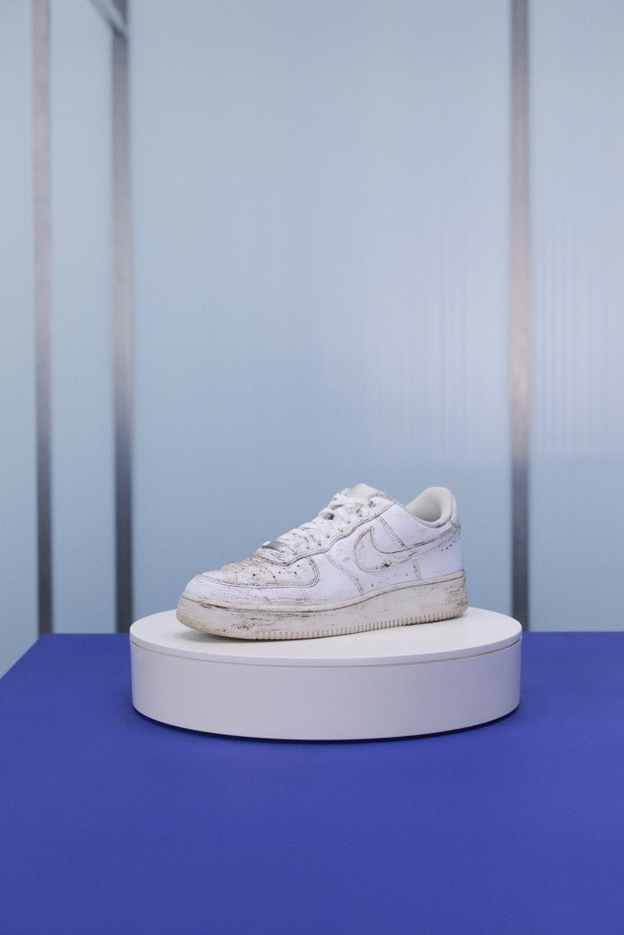 Vanhentunut valkoinen Nike Air Force 1 -kengät asetettuna pyöreälle jalustalle sinistä taustaa vasten, korostaen niiden naarmuista ja likaisesta ulkonäöstä.