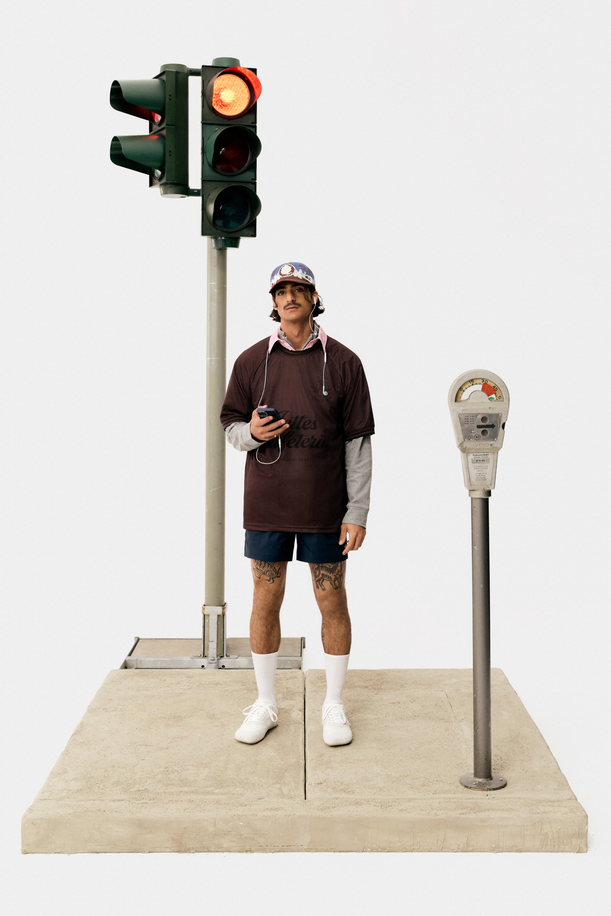 A young man stands at a crosswalk, holding a phone, with a traffic light showing red and a parking meter beside him.