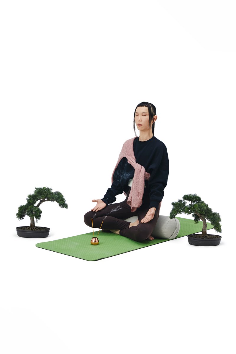 Person meditating on a green mat, flanked by two bonsai trees. A small incense holder emits smoke. The setting emphasizes tranquility and mindfulness.