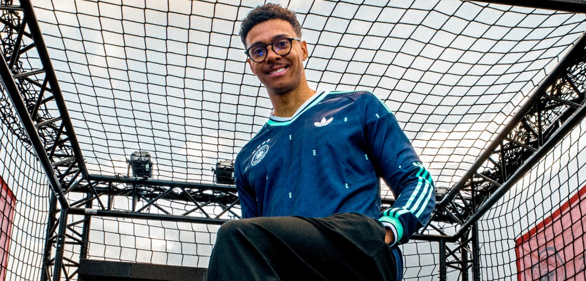 Junger Mann mit Brille, der ein blaues Adidas-Trikot trägt, steht in einer metallgerahmten Netzstruktur und lächelt in die Kamera.