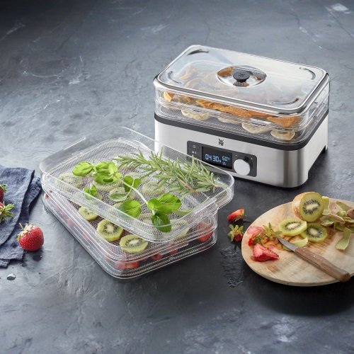 Przezroczyste prostokątne pojemniki na jedzenie są ułożone w stos, poniżej znajduje się cyfrowa jednostka sterująca. Pojemniki mają otwory wentylacyjne i są wypełnione owocami oraz ziołami.