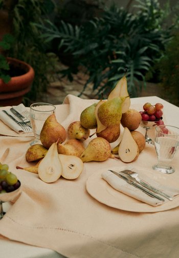 Birnen, einige ganze und geschnittene, arrangiert auf einem beigen Tischtuch mit klaren Gläsern, Besteck auf Servietten und Schüsseln mit Trauben auf einem Gartentisch.