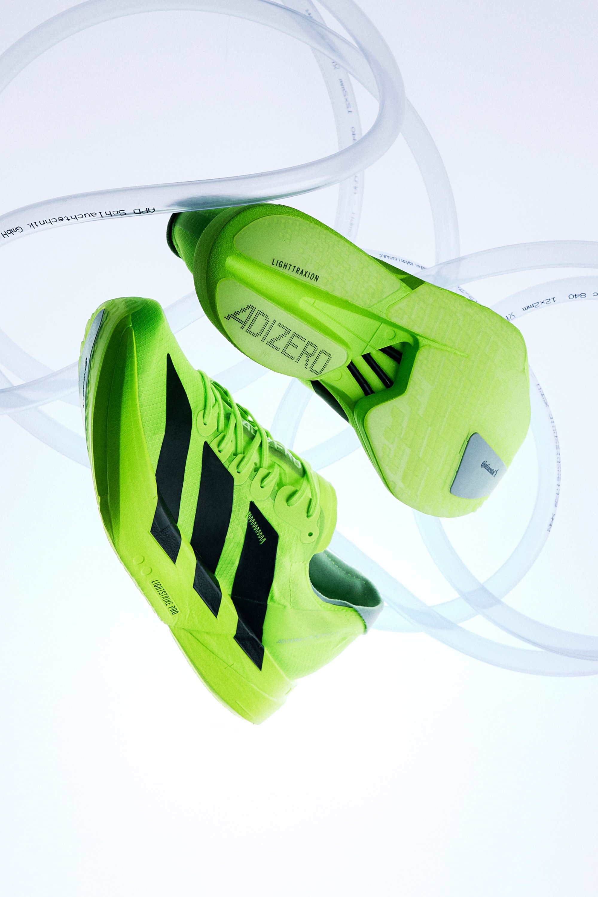Baskets de course Adidas vert vif avec des rayures noires suspendues à côté de tubes translucides enroulés sur un fond blanc.
