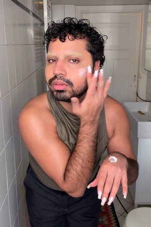 Applying cream to the face, the person has curly black hair, light-colored eyebrows, and long, shiny nails. They're wearing a dark top.