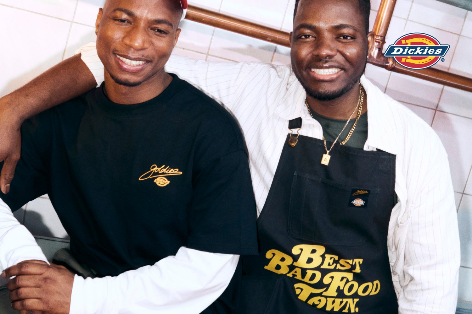 Zwei lächelnde Männer posieren zusammen in einer Küche. Einer trägt ein schwarzes Shirt mit dem Text "goldie", der andere hat eine Schürze an, auf der steht "Best Bad Food Town."