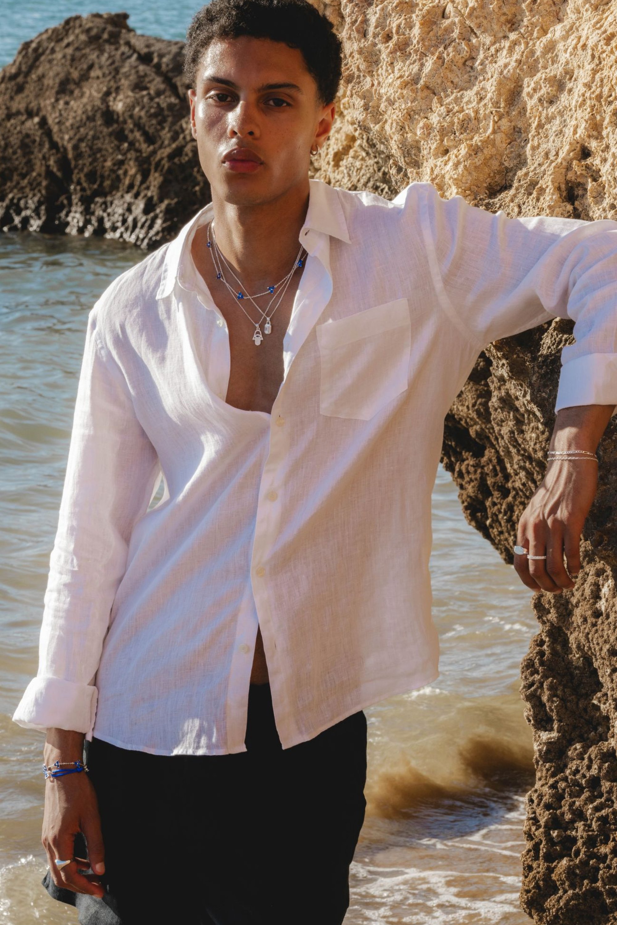 Camisa de lino blanca con botones y un bolsillo en el pecho, llevada abierta sobre pantalones negros. La joyería de plata incluye collares en capas y pulseras.