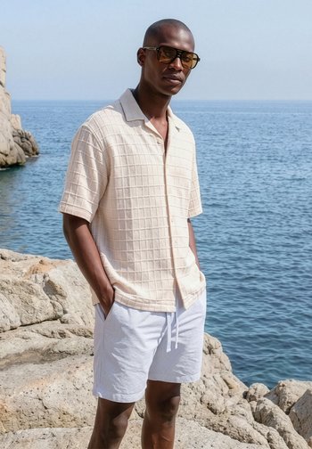 Homme portant une chemise beige à manches courtes et un short blanc se tient sur une côte rocheuse avec l’océan et un ciel dégagé en arrière-plan.