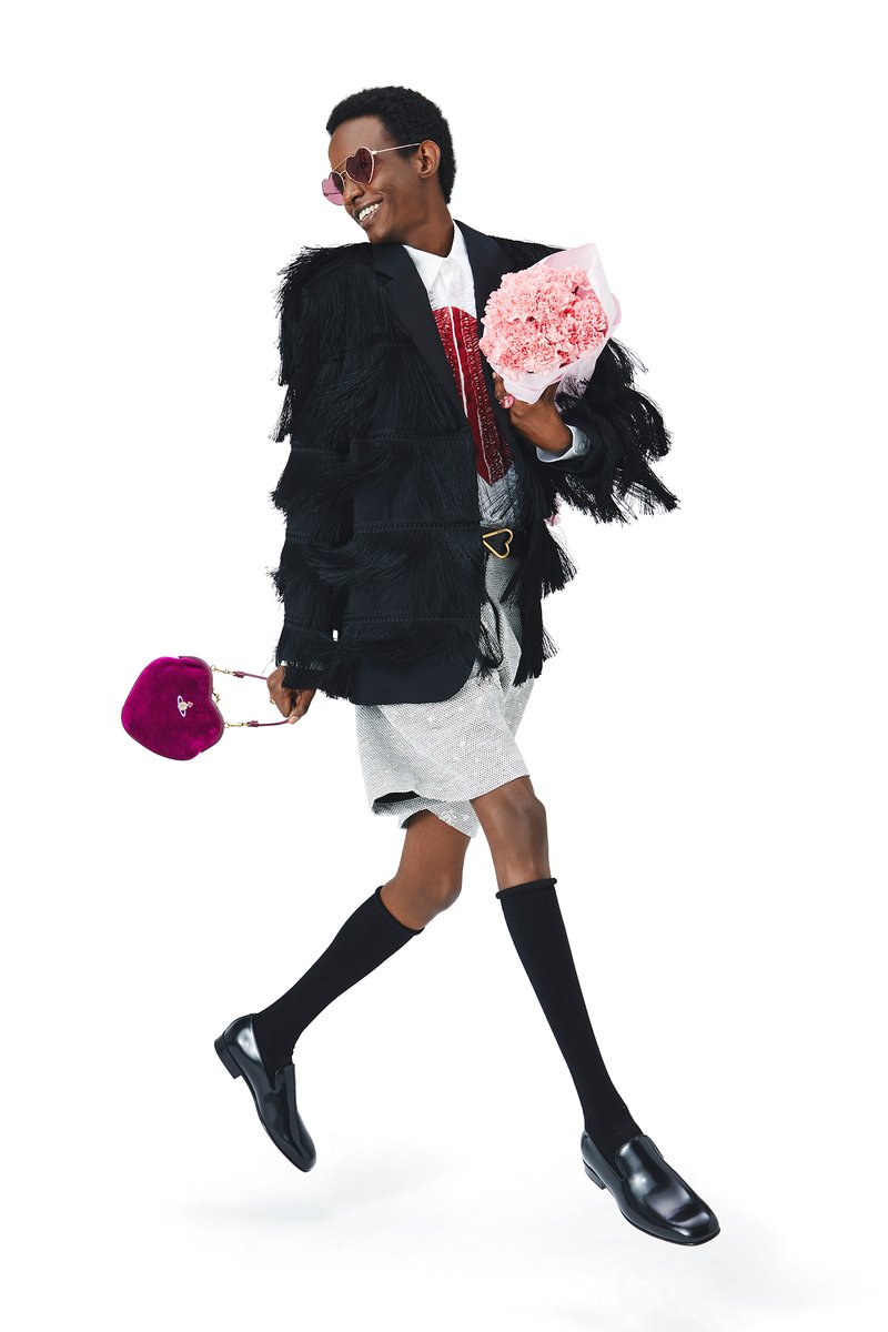 Una persona sonriente con una chaqueta negra con flecos y una falda plateada camina, sosteniendo un ramo de flores rosas y un bolso en forma de corazón rosa.