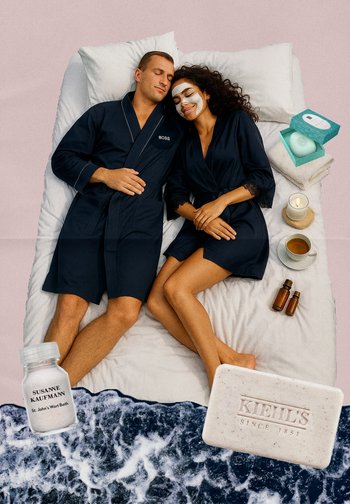 Deux personnes en peignoirs bleus marine sont allongées sur un lit blanc. Autour d'eux se trouvent des produits de soin de la peau et une bougie, avec un fond décoratif apaisant.