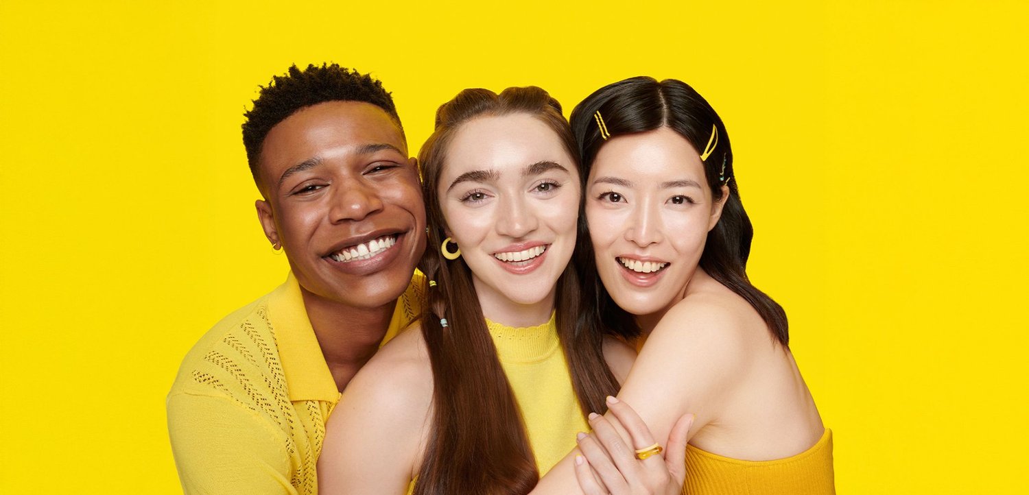 Drei Personen in gelber Kleidung lächeln vor einem leuchtend gelben Hintergrund. Accessoires sind Haarklammern und Ohrringe.