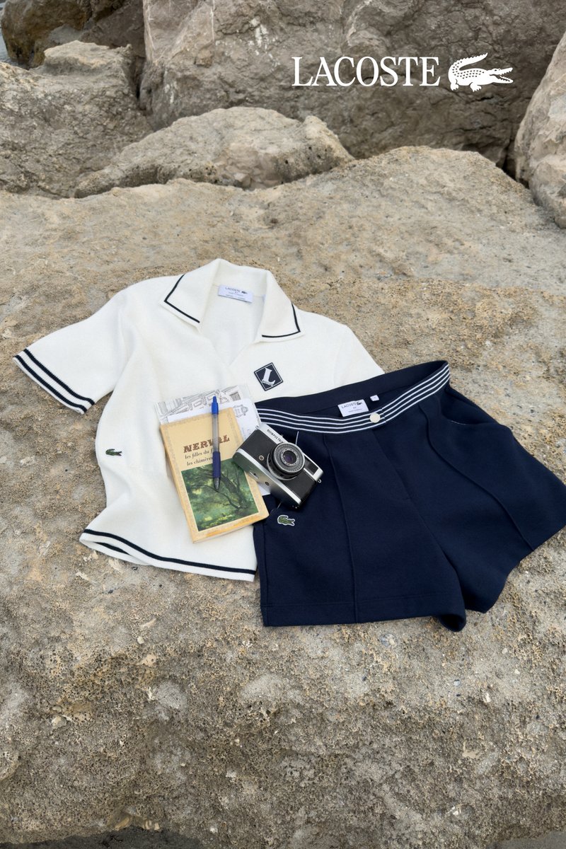 Weißes Lacoste Kurzarmhemd und marineblaue Shorts auf Felsen mit Buch, Stift, Vintage-Kamera und Lacoste-Logo im Hintergrund.