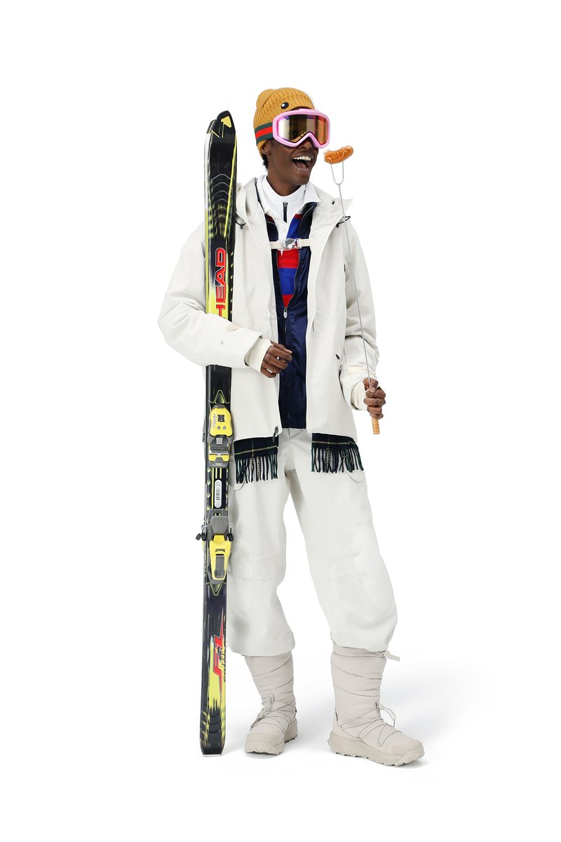 Een lachende persoon in wintersportoutfit, die skistokken en ski's vasthoudt, met roze goggles en een gezellige muts, poseert in een speelse houding.
