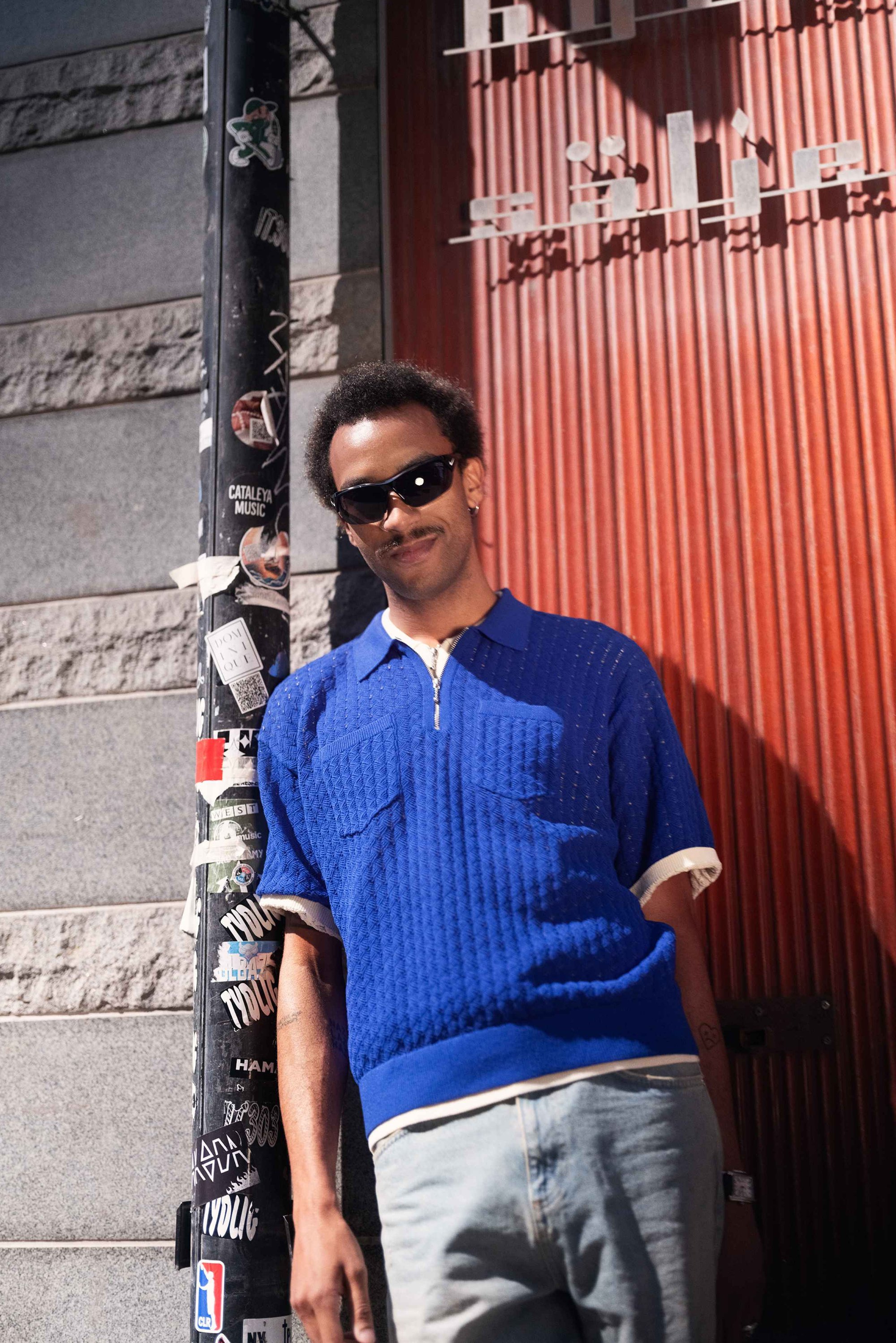 Blauwe gebreide polo met ritssluiting en twee voorzakken, gecombineerd met lichte jeans. Zwarte zonnebril maakt de look compleet.