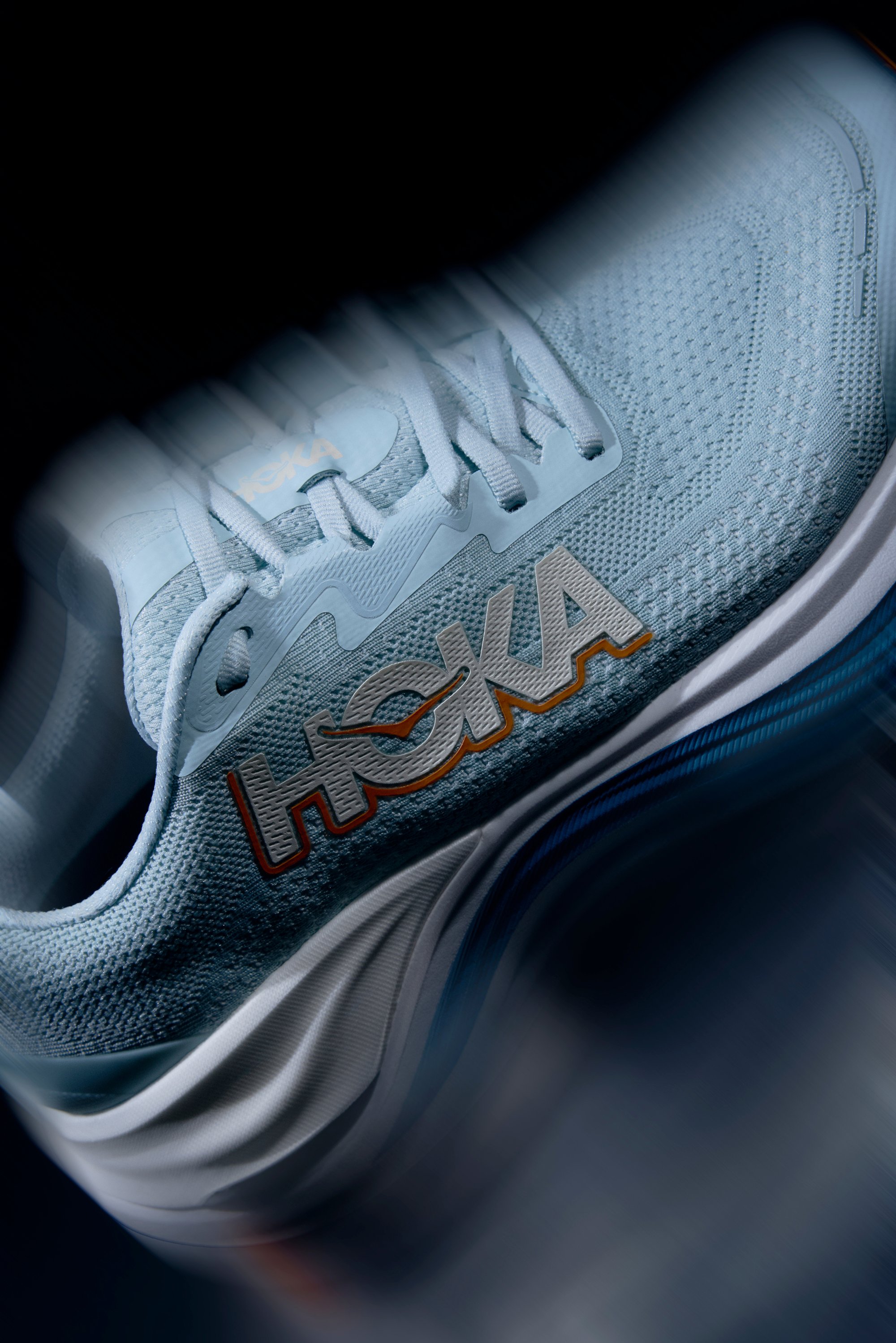 Světle modrá sportovní bota s texturovaným síťovaným svrchním materiálem, bílou mezipodešví a vícolorovým logem "HOKA" na boku. Obsahuje zaoblený design špičky.
