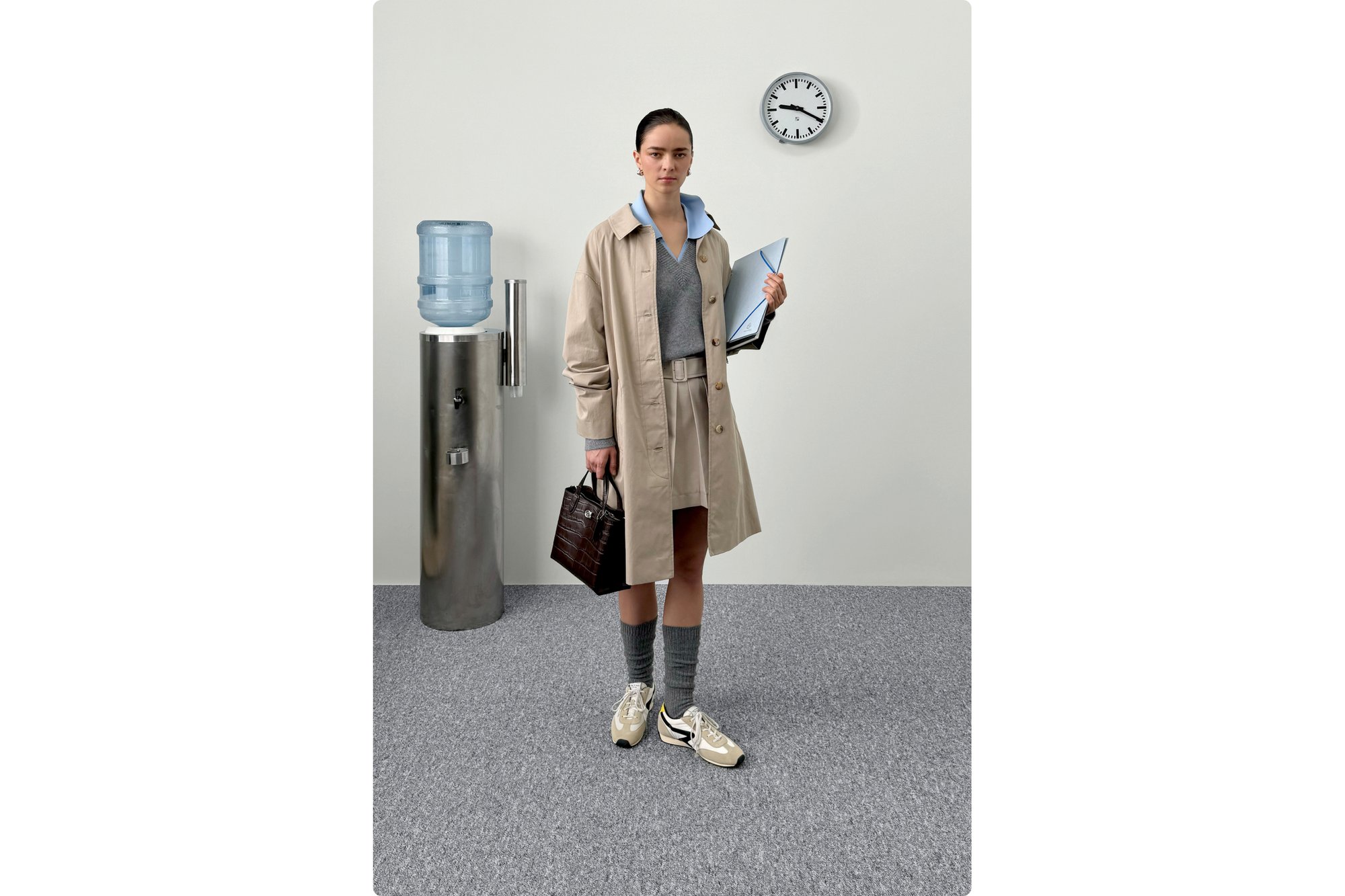 Una joven con un abrigo trenzado beige está de pie junto a un enfriador de agua, sosteniendo una carpeta y un bolso. Un reloj es visible en la pared.