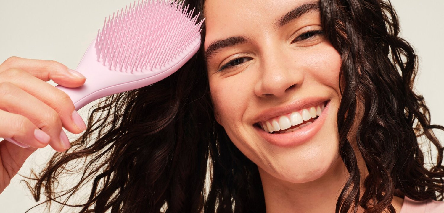 Giovane donna con capelli ricci e scuri sorride mentre si pettina con una spazzola rosa.