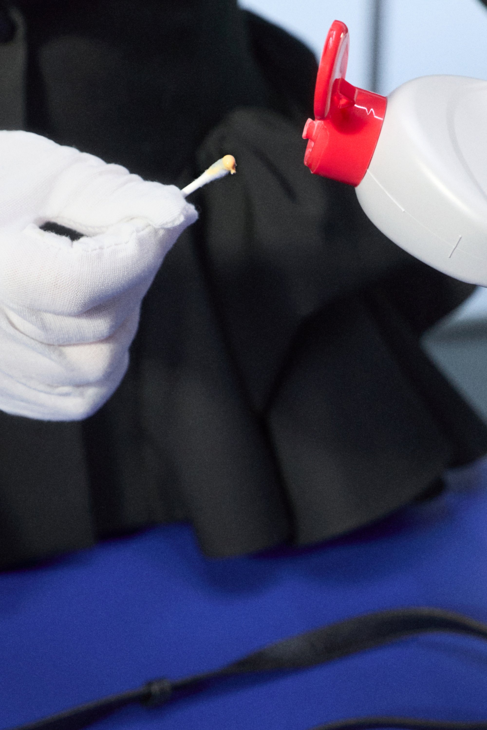 Una mano guantata tiene un fiammifero acceso vicino all'ugello di una bottiglia spray, con uno sfondo scuro e una superficie blu visibile, insieme a una tracolla.